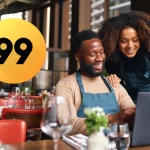 Duas pessoas sorrindo enquanto configuram o cardápio do restaurante no laptop, com o logo da 99Food ao lado. Imagem representa empresários do setor de food service iniciando suas vendas na 99Food com apoio de um sistema de gestão.