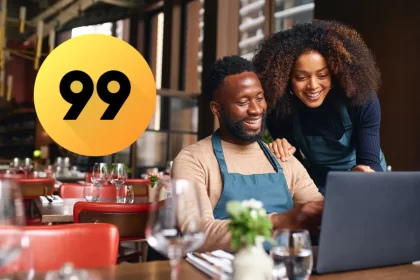 Duas pessoas sorrindo enquanto configuram o cardápio do restaurante no laptop, com o logo da 99Food ao lado. Imagem representa empresários do setor de food service iniciando suas vendas na 99Food com apoio de um sistema de gestão.