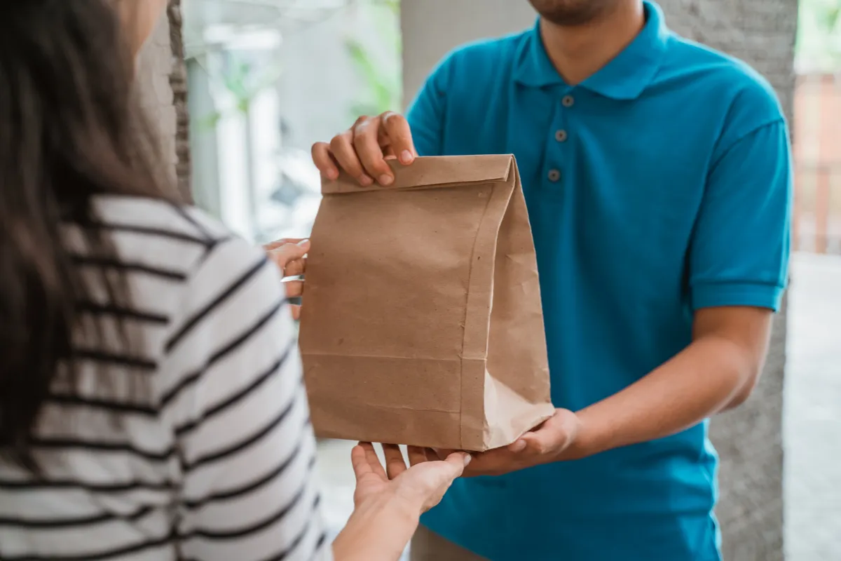 Entregador com camisa azul entrega embalagem de papel pardo para cliente. Foto representa exigências sanitárias no delivery, como embalagem lacrada e transporte seguro.