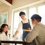 Garçom sorridente servindo bebida para casal em restaurante durante o dia, ilustrando atendimento cordial em estabelecimentos que costumam sugerir os 10% de taxa de serviço.