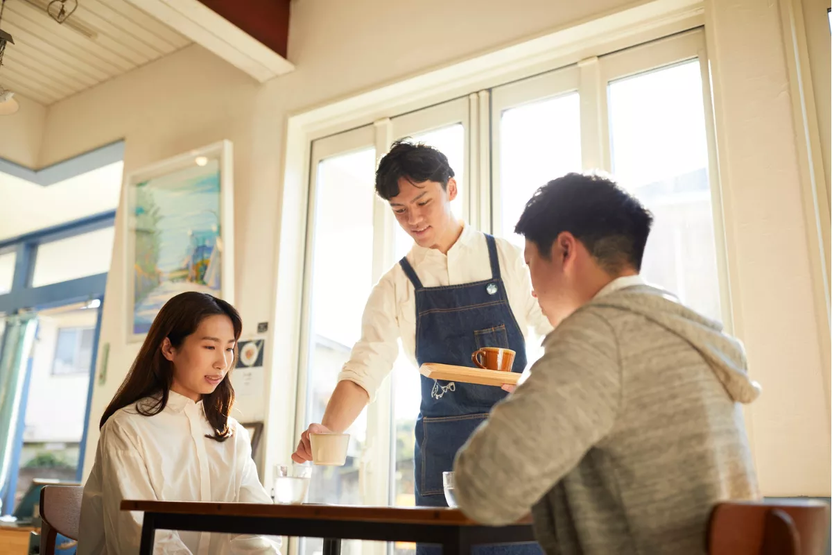 Garçom sorridente servindo bebida para casal em restaurante durante o dia, ilustrando atendimento cordial em estabelecimentos que costumam sugerir os 10% de taxa de serviço.