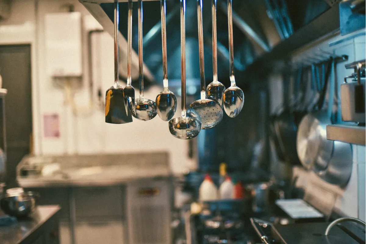Utensílios de cozinha pendurados de forma organizada em ambiente limpo. Representa a exigência de equipamentos em bom estado e higiene adequada na área de preparo.

