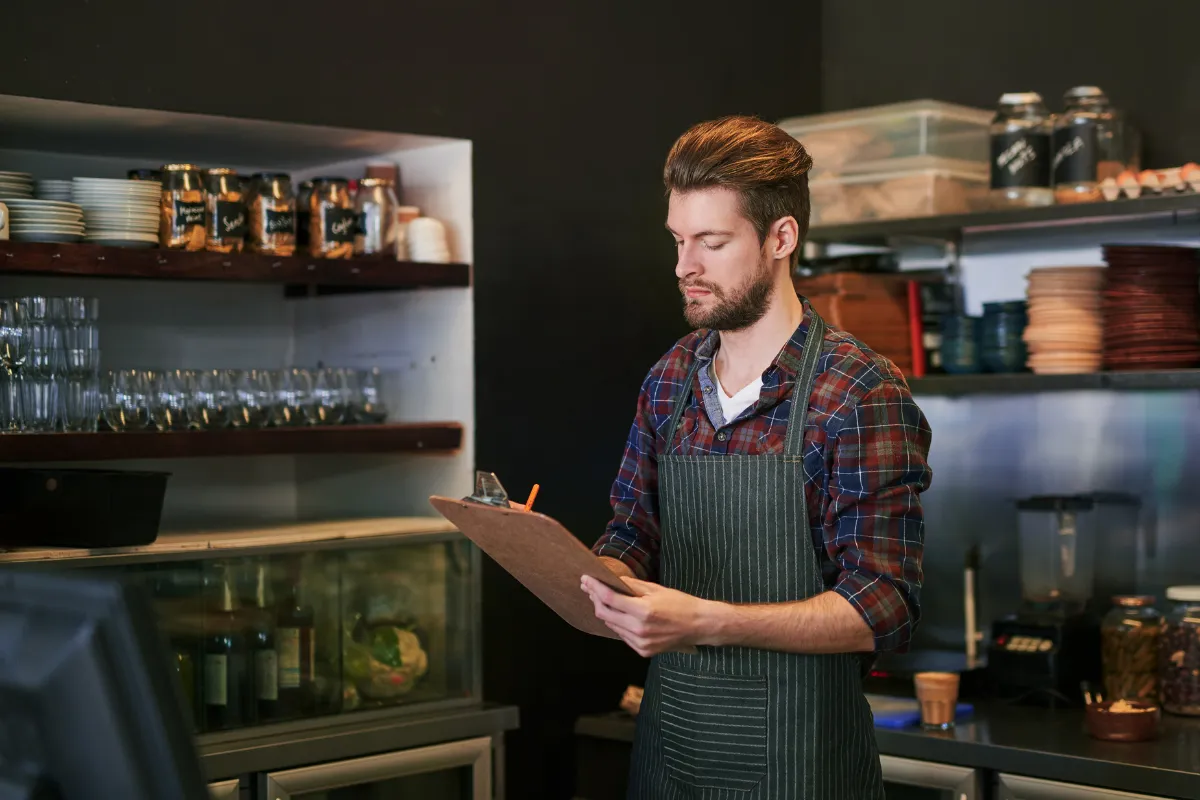 Dono de restaurante revisando checklist de operação com prancheta em mãos, simbolizando o controle de processos e a organização necessária na fase inicial do negócio.