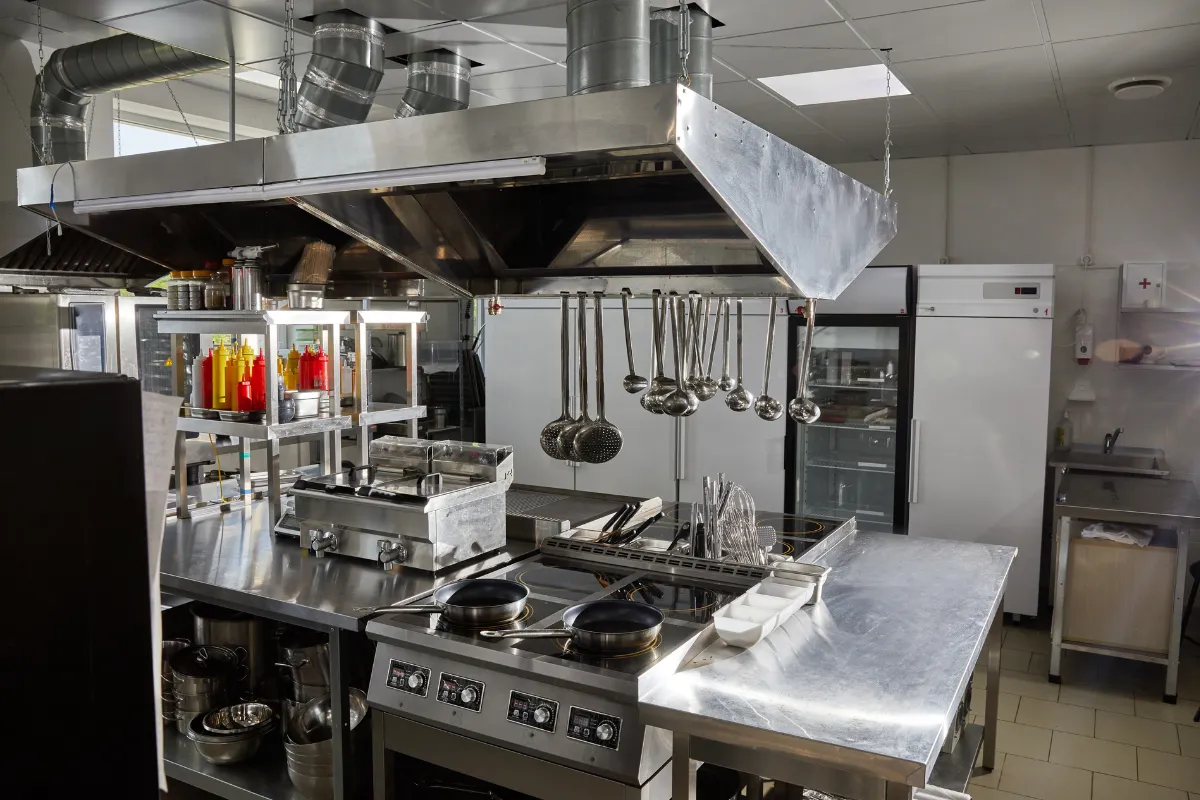 Cozinha industrial organizada com equipamentos inox, exaustores e iluminação adequada. Imagem mostra padrão exigido de estrutura física e higiene no ambiente de preparo.


