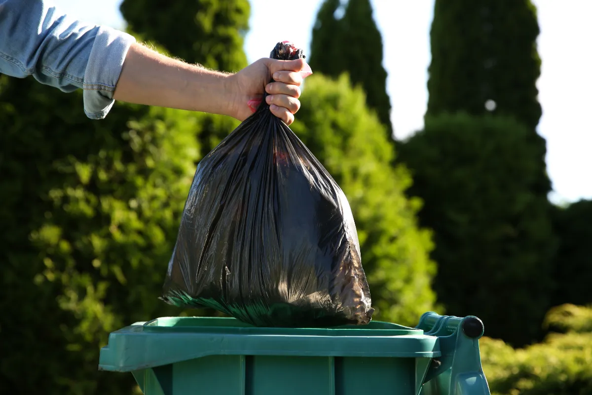 Pessoa descartando saco de lixo preto em lixeira externa. Ilustra a correta gestão de resíduos exigida pela Vigilância Sanitária para evitar contaminação e pragas.


