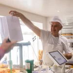 Funcionário de padaria entregando pedido em embalagem para viagem a cliente no balcão, representando modelo Grab and Go com atendimento rápido e prático.