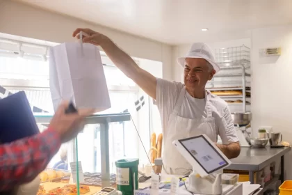 Funcionário de padaria entregando pedido em embalagem para viagem a cliente no balcão, representando modelo Grab and Go com atendimento rápido e prático.