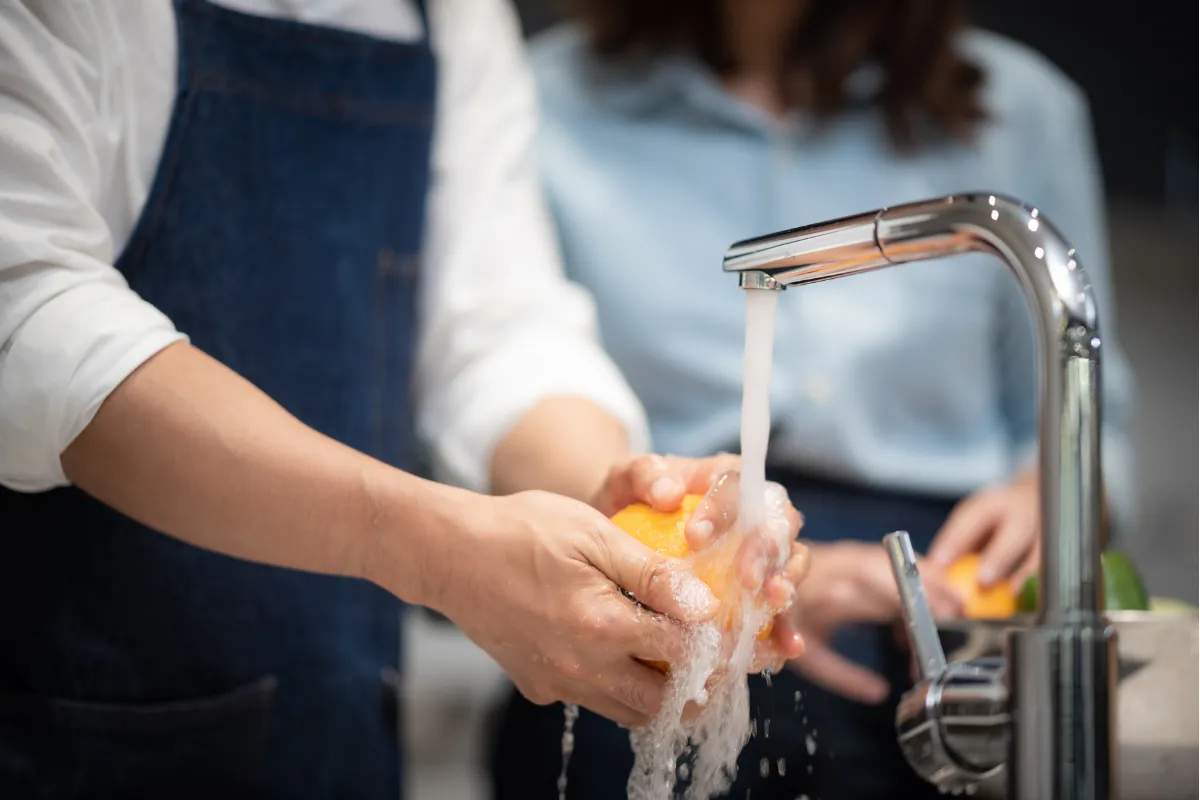 Profissional de cozinha lavando fruta sob água corrente. Representa a prática obrigatória de higiene pessoal e manipulação segura dos alimentos.