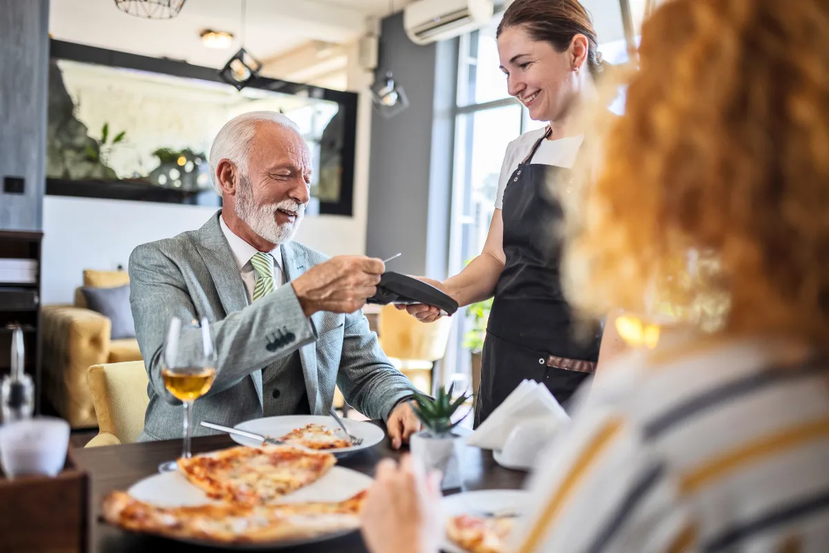Pai mais velho sorrindo ao pagar a conta em um restaurante elegante, sendo atendido por uma garçonete, ilustrando celebrações familiares e o impacto positivo de promoções de Dia dos Pais no setor de food service.