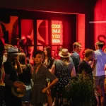 Pessoas reunidas em frente a bar com luz vermelha e letreiro de neon, ilustrando bares movimentados com atmosfera noturna e urbana.