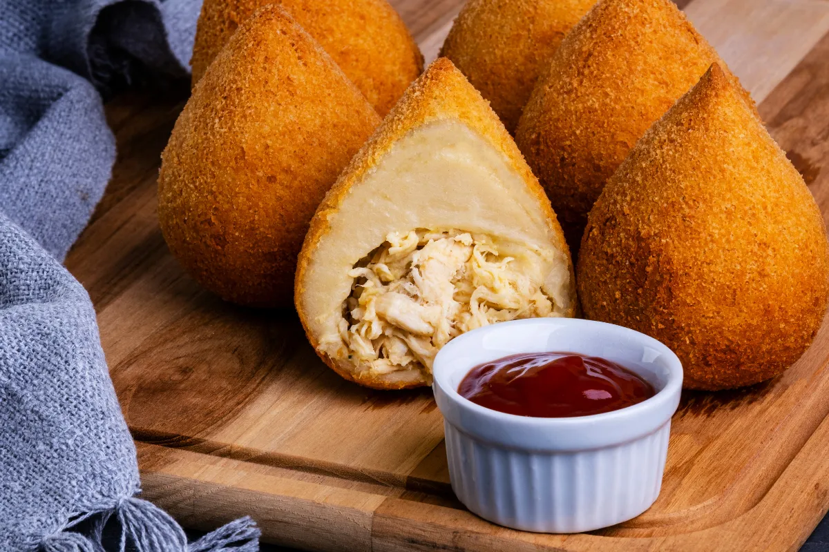 Coxinhas crocantes e recheadas com frango desfiado, acompanhadas de molho, ilustrando o carro-chefe clássico de muitas lojas de salgados no Brasil.

