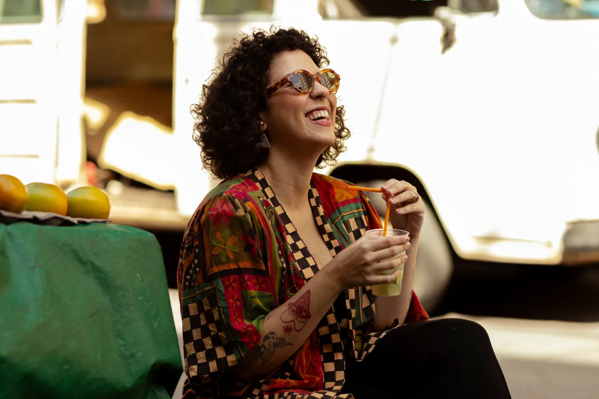Mulher sorrindo enquanto toma uma caipirinha na rua, em frente a uma barraca com frutas, sugerindo bares ao ar livre ou com temática brasileira.
