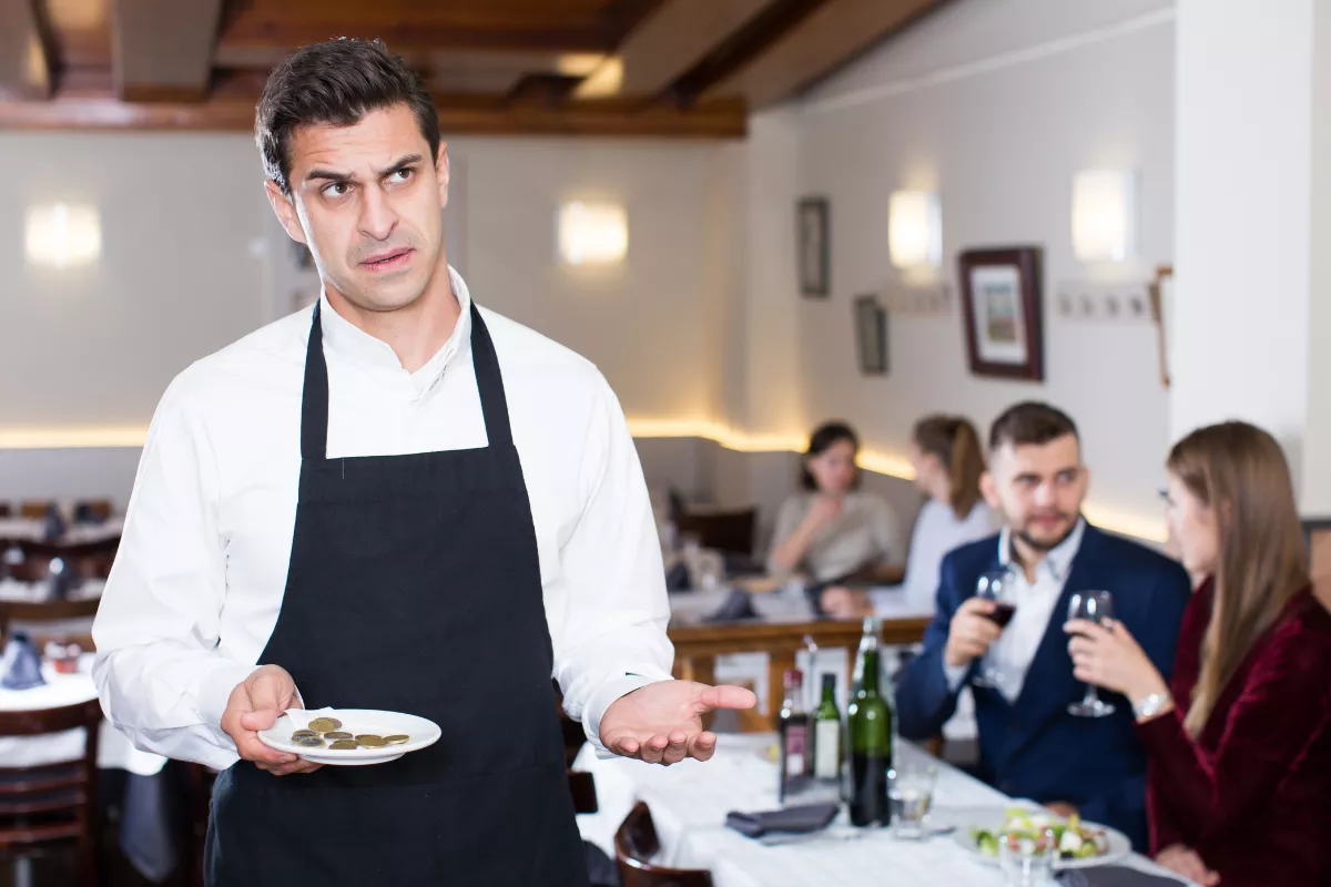 Garçom com expressão de insatisfação segurando prato com moedas, em restaurante, sugerindo frustração com gorjeta baixa ou ausência da taxa de serviço opcional.