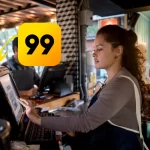 Atendente de restaurante usando sistema PDV em frente de caixa, com destaque para o ícone da 99Food, simbolizando integração entre o sistema e a plataforma de delivery.