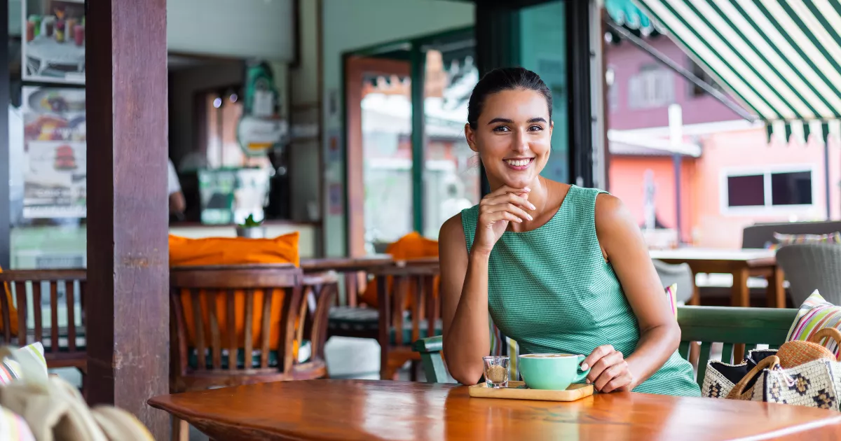 Mulher sorridente sentada à mesa de um restaurante pequeno, com uma xícara sobre a mesa.