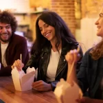 Grupo de amigos se divertindo enquanto comem comida oriental em embalagens para viagem — imagem que reforça nomes criativos e urbanos para deliverys.