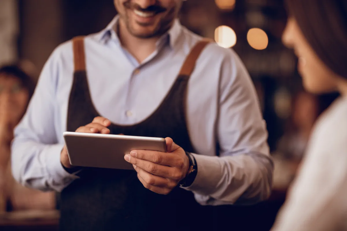 Garçom atendendo cliente com tablet em mãos, demonstrando como o aplicativo para garçom agiliza o registro e envio de pedidos no salão.