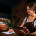 Mulher gestora de padaria analisando informações fiscais e anotando dados com tablet e caderno, em ambiente de produção de pães artesanais.