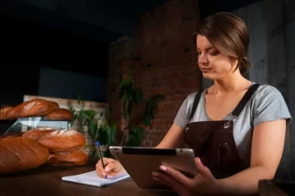 Mulher gestora de padaria analisando informações fiscais e anotando dados com tablet e caderno, em ambiente de produção de pães artesanais.