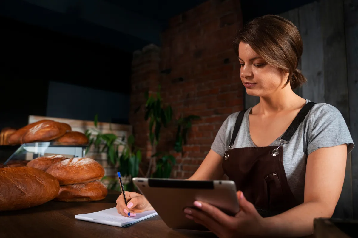 Mulher gestora de padaria analisando informações fiscais e anotando dados com tablet e caderno, em ambiente de produção de pães artesanais.