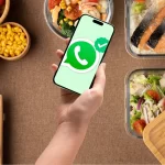 Mão segurando um celular com o ícone do WhatsApp em destaque, cercado por marmitas saudáveis e bem apresentadas.