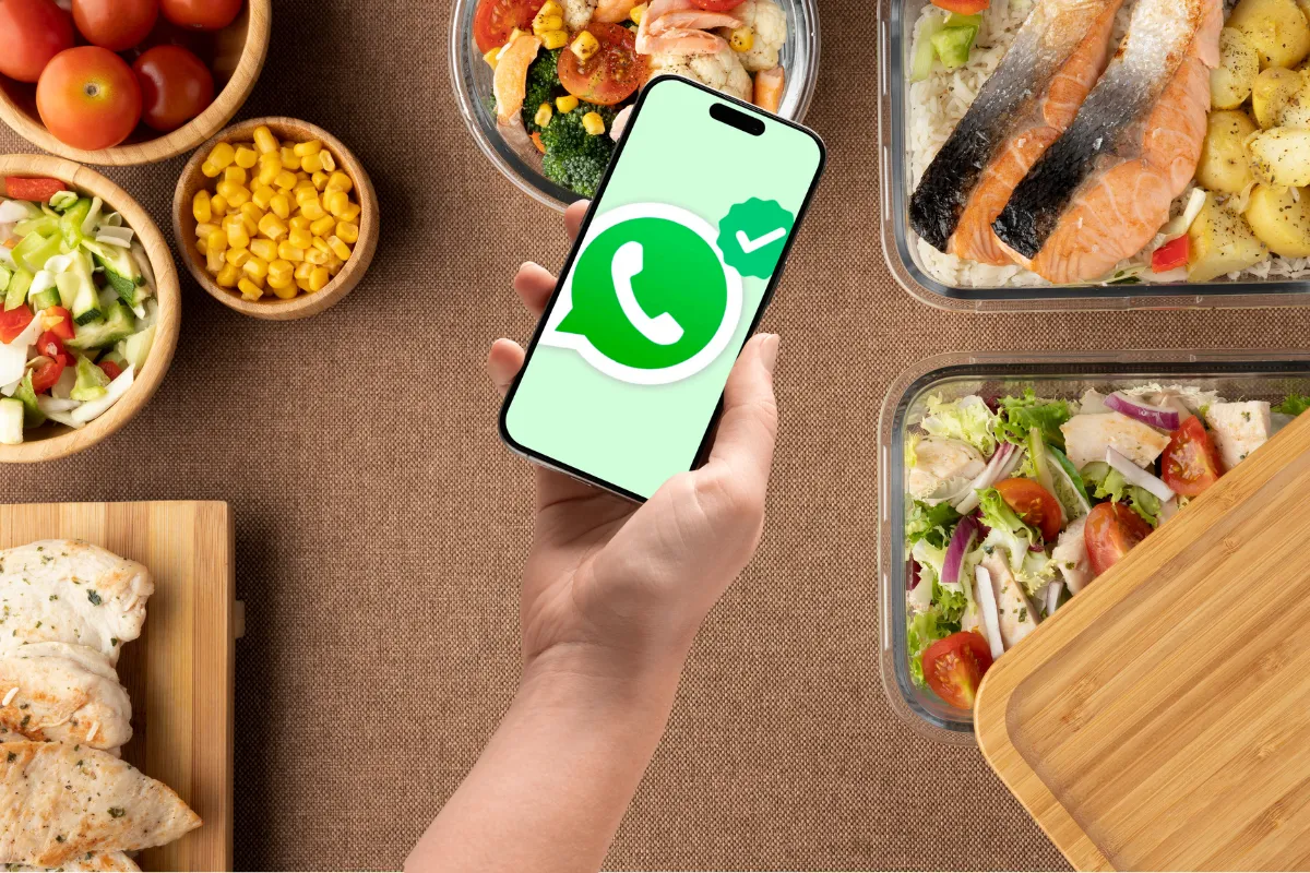Mão segurando um celular com o ícone do WhatsApp em destaque, cercado por marmitas saudáveis e bem apresentadas.