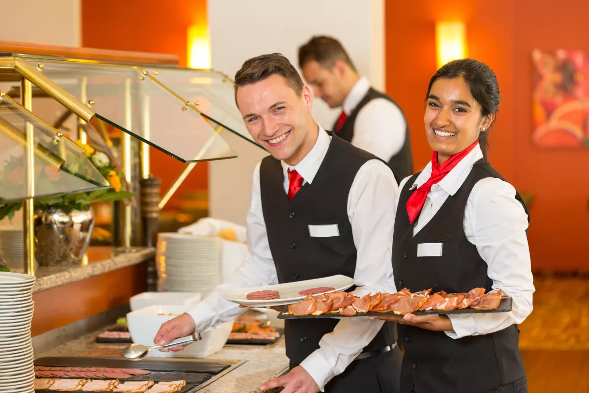 Dois garçons sorridentes servindo alimentos em um buffet de restaurante, representando a importância do dimensionamento correto da equipe de atendimento.