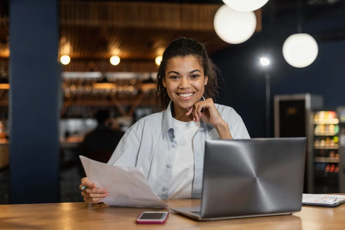 Empreendedora de restaurante sorrindo enquanto organiza documentos fiscais e trabalha com notebook, demonstrando controle e gestão eficiente do negócio.