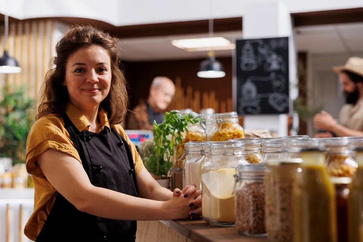  Proprietária de restaurante em frente ao balcão com potes de ingredientes, simbolizando controle de estoque e compras estratégicas conforme a sazonalidade da demanda.