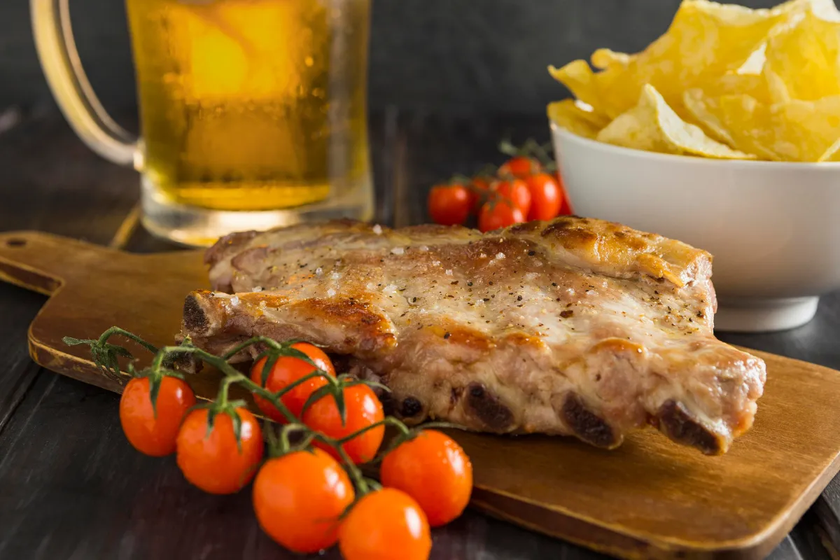 Costela suína assada com batatas chips e cerveja ao fundo, prato rústico e saboroso, perfeito para surpreender pais gourmet no almoço de Dia dos Pais.