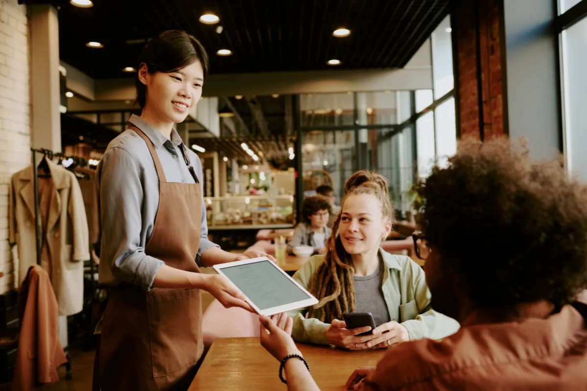 Garçonete apresentando tablet a clientes em restaurante moderno, evidenciando a interação entre o app para garçom e o cardápio digital.
