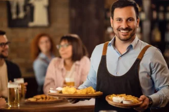 Atendente leva duas porções de petiscos a clientes sentados em uma petiscaria, com copos de cerveja sobre a mesa.