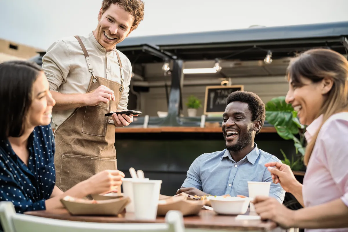 Clientes sorrindo enquanto são atendidos por garçom em food truck moderno — cenário descontraído que combina com nomes leves e engraçados para restaurantes.