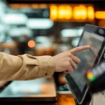 Pessoa tocando na tela de um terminal de autoatendimento para fazer pedido e pagamento em restaurante.
