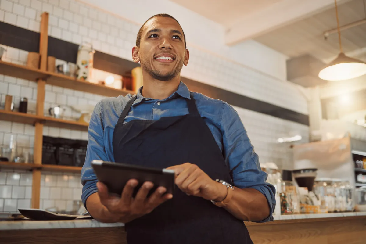 Dono de marmitaria sorrindo enquanto utiliza um tablet para gerenciar pedidos e atendimento, em ambiente de restaurante.