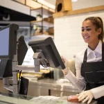 Atendente sorridente operando um sistema de PDV moderno em um balcão de atendimento, com duas telas integradas para registrar pedidos e gerenciar vendas em tempo real.