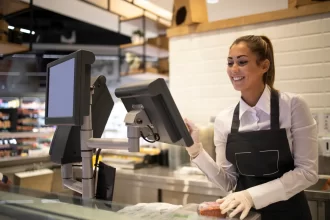 Atendente sorridente operando um sistema de PDV moderno em um balcão de atendimento, com duas telas integradas para registrar pedidos e gerenciar vendas em tempo real.