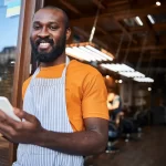 Garçom sorridente com avental usando smartphone na porta de um restaurante, simbolizando a mobilidade e autonomia com app de atendimento.