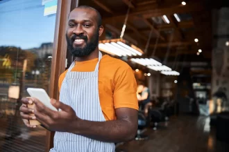 Garçom sorridente com avental usando smartphone na porta de um restaurante, simbolizando a mobilidade e autonomia com app de atendimento.