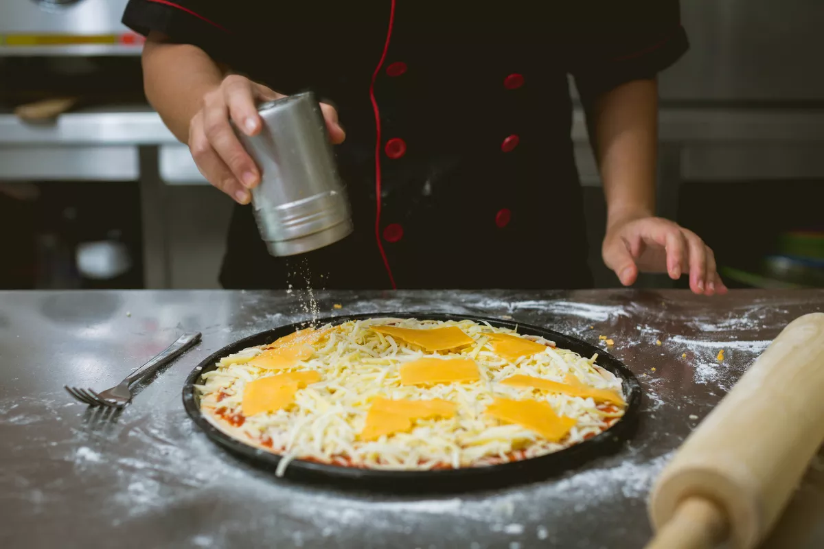 Mãos de um chef polvilhando queijo ralado sobre uma pizza coberta com queijo fatiado em uma assadeira, destacando o processo de condimentação antes de assar.