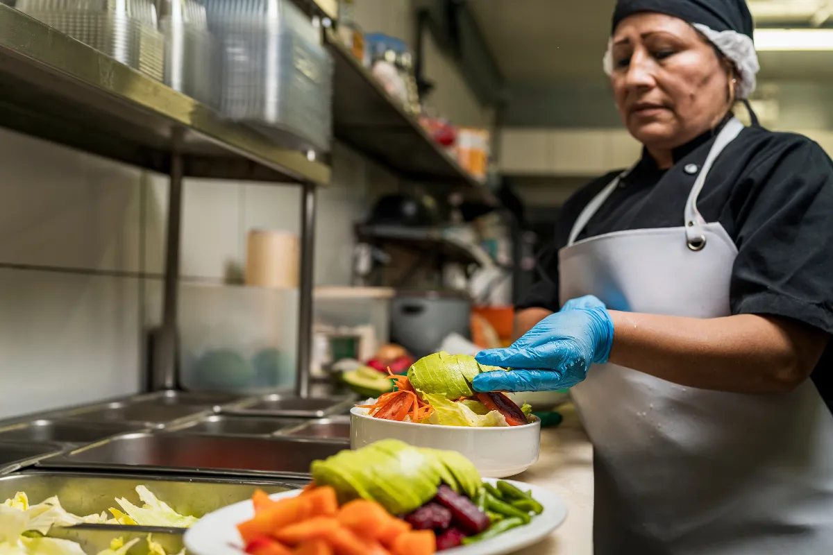 Auxiliar de cozinha preparando salada fresca com luvas, representando o mise en place e o suporte essencial ao trabalho do cozinheiro.
