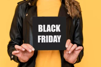 Mulher de jaqueta de couro preta e blusa amarela segura um cartão preto escrito Black Friday em letras brancas, diante de um fundo amarelo, marcando a data de promoções