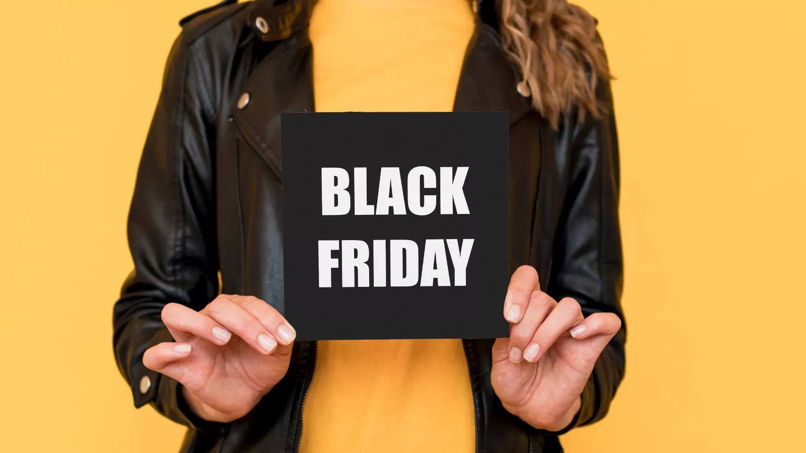 Mulher de jaqueta de couro preta e blusa amarela segura um cartão preto escrito Black Friday em letras brancas, diante de um fundo amarelo, marcando a data de promoções