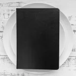Uma prancheta preta em branco, colocada sobre um prato de porcelana branca, com um garfo e uma faca ao lado. A imagem simboliza a importância do planejamento e precificação estratégica do cardápio para a Black Friday.