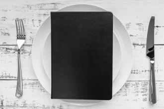 Uma prancheta preta em branco, colocada sobre um prato de porcelana branca, com um garfo e uma faca ao lado. A imagem simboliza a importância do planejamento e precificação estratégica do cardápio para a Black Friday.