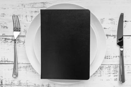 Uma prancheta preta em branco, colocada sobre um prato de porcelana branca, com um garfo e uma faca ao lado. A imagem simboliza a importância do planejamento e precificação estratégica do cardápio para a Black Friday.
