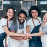 Equipe de restaurante unida e sorridente, com mãos juntas, demonstrando espírito de equipe e colaboração entre diferentes cargos da operação.