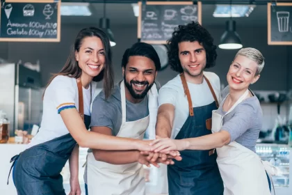 Equipe de restaurante unida e sorridente, com mãos juntas, demonstrando espírito de equipe e colaboração entre diferentes cargos da operação.