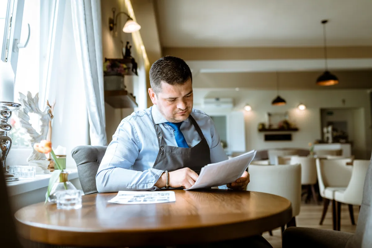 Dono de restaurante analisando documentos financeiros à mesa do salão. Representa o desafio de calcular corretamente a alíquota do Simples Nacional para restaurantes.