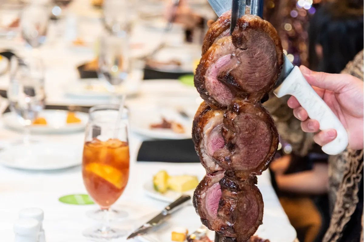 Carne assada sendo servida no espeto em churrascaria, com mesa posta ao fundo, representando a experiência do rodízio que impacta no faturamento do setor.